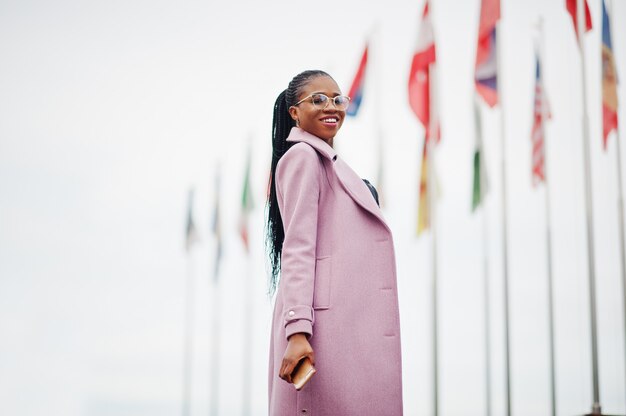 젊은 세련 된 아름 다운 아프리카 계 미국인 여자 거리에서 손에 휴대 전화와 함께 세계의 다른 나라의 국기에 대 한 패션 복장 코트를 입고.