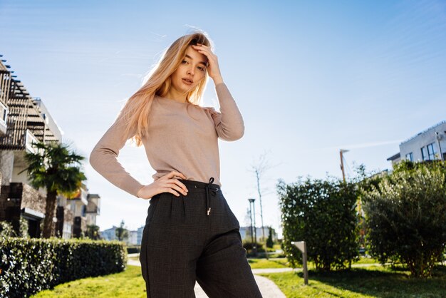 Young stylish blonde girl posing outdoors, in the sun, looking at the camera