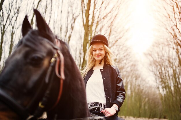 Young stylish blonde girl riding on horses at autumn forest