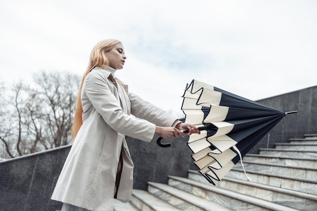 Young stylish blonde girl in a trench coat opens an umbrella climbing the stairs