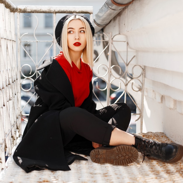 Photo young stylish blonde woman with red lips in a fashionable coat with a black beret in an elegant red blouse in leather boots is sitting near a vintage metal railing on a sunny autumn day