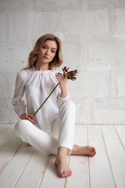 A young stylish blueeyed lady in a white pantsuit is sitting on the floor in a romantic mood