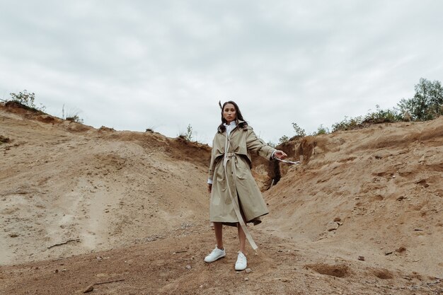 Giovane ed elegante ragazza bruna in posa dinamicamente in un trench beige nella cava di sabbia.