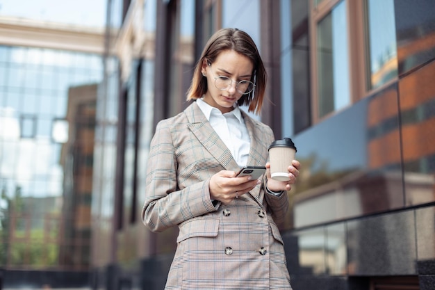 Young stylish business woman using smartphone and holding coffee cup in city