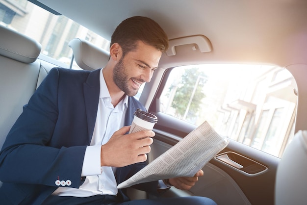 Young stylish businessman with newspaper drinking coffee on backseat of car and laughs