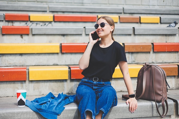 Young stylish businesswoman sitting on the steps and holding a phone and a cup of coffee