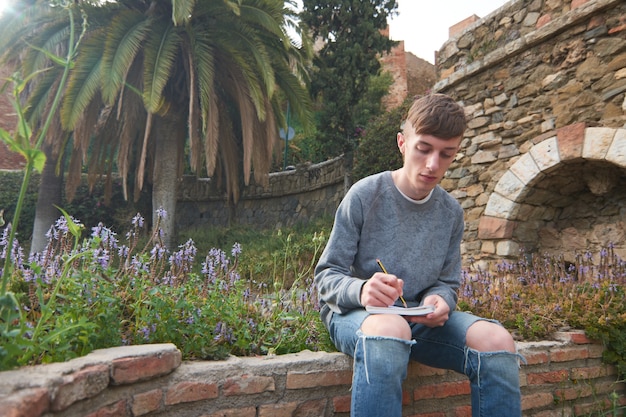 Молодой стильный кавказский парень сидит в саду и делает домашнее задание