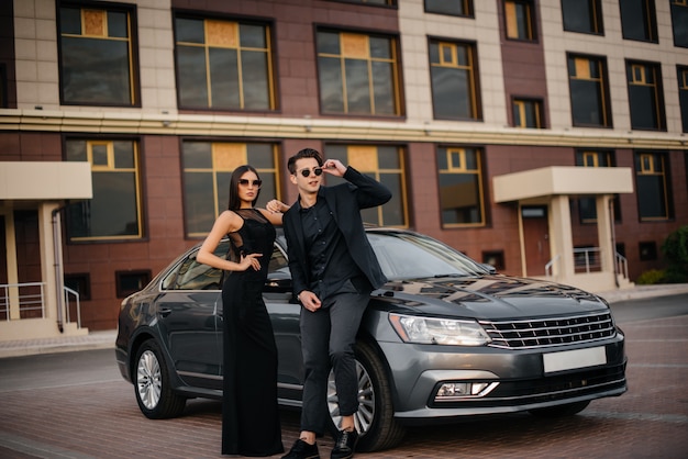 A young stylish couple in black stands near the car at sunset. Fashion and style