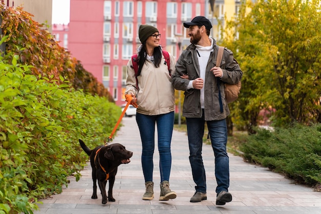 通りで犬と一緒に歩いている若いスタイリッシュなカップル。男と女はお互いを見ながら一緒に幸せ。ラブラドール犬種、秋の季節、カジュアルなスタイル