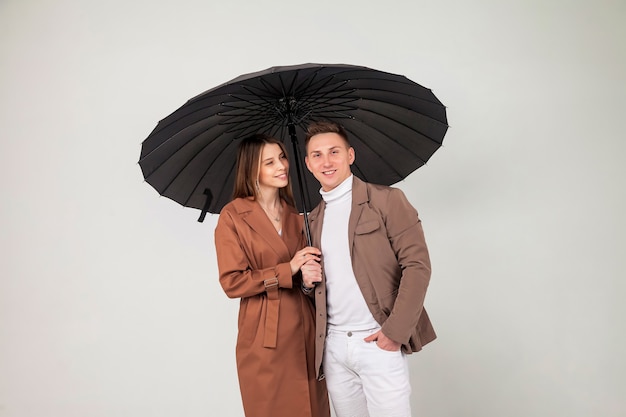 Молодая стильная пара с черным зонтиком показывает любовные эмоции. Портрет милых эмоционально людей в осенней одежде, стоящих под зонтиком, глядя на светло-сером фоне. Скопируйте место для сайта