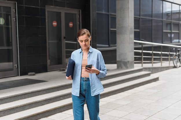 Young stylish female businesswoman leaves the office center and looks at the phone. The concept of feminism and women's business.