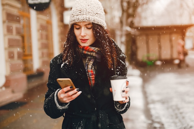 young and stylish girl in black coat and white hat drinking coffee in winter city