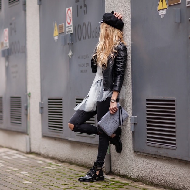 Young stylish girl in a black leather jacket with a gray handbag in her hands looks away