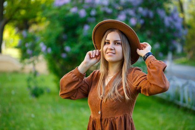 Young stylish girl in a hat near the lush bush of pink lilac in the garden. The girl enjoys the smell of lilac. Warm summer day in the garden young woman with a smile on her face