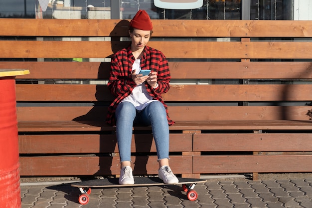 Young stylish girl skater chatting in smartphone outdoors sitting on bench after longboard riding