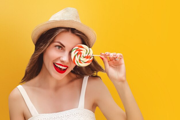 Giovane ragazza alla moda in un cappello di paglia con una lecca-lecca arcobaleno su uno sfondo giallo