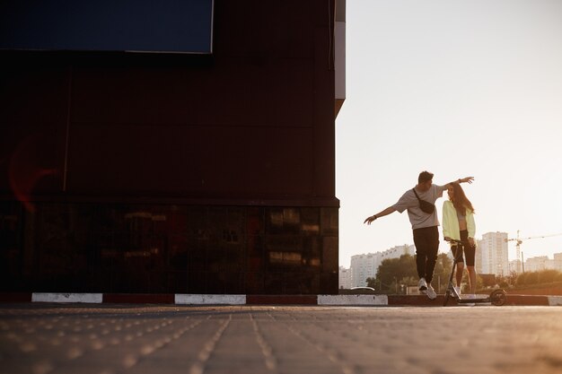 写真 若いスタイリッシュな男と女が夕日の建物の隣に駐車するために広場を一緒に歩いています。