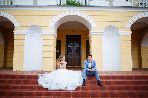 Молодой стильный парень в костюме жениха и красивая девушка невесты в белом платье с поездом гуляют по парку в день их свадьбы