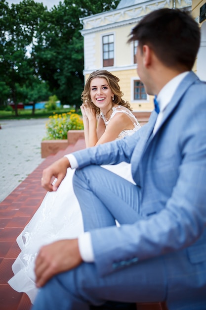 Молодой стильный парень в костюме жениха и красивая девушка невесты в белом платье с поездом гуляют по парку в день их свадьбы