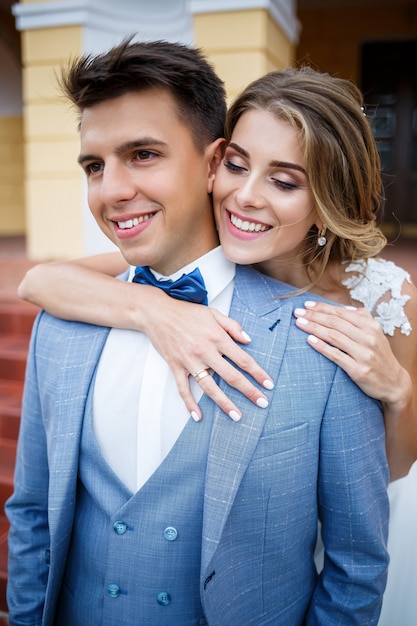 Young stylish guy in the costume of the groom and the bride beautiful girl in a white dress with a train walking on the background of a large house with columns on their wedding day