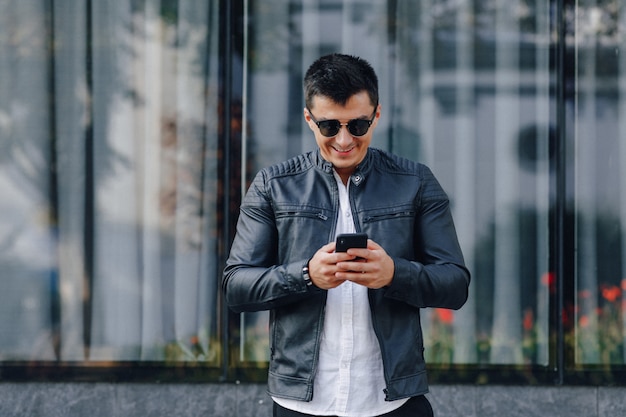 Young stylish guy in glasses in black leather jacket with phone on glass background