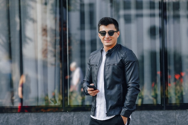 Young stylish guy in glasses in black leather jacket with phone on glass background