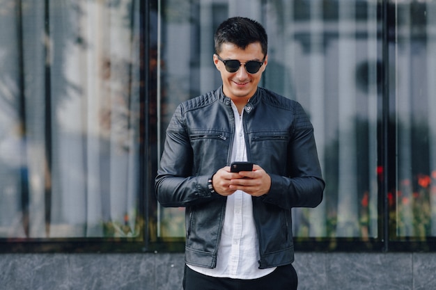 Young stylish guy in glasses in black leather jacket with phone on glass background