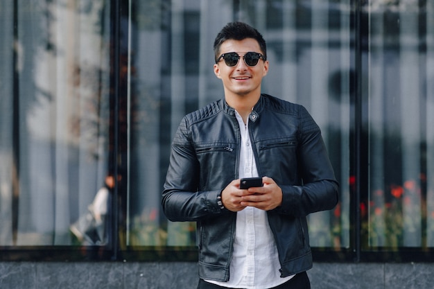 Giovane ragazzo elegante con gli occhiali in giacca di pelle nera con telefono