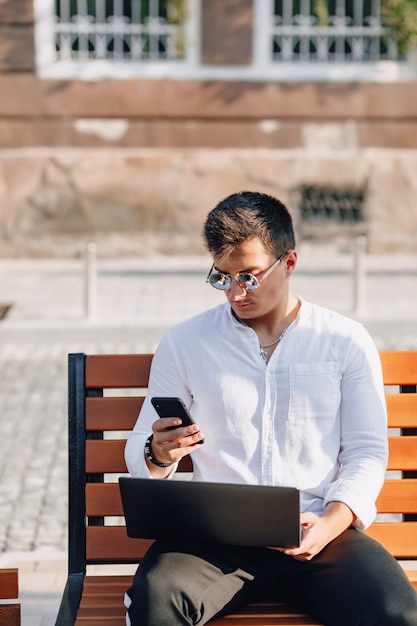 Young stylish guy in shirt with phone and notebook on bench on sunny warm day outdoors, freelance