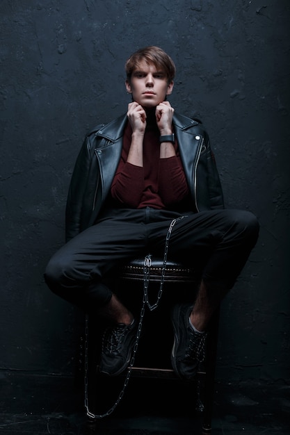 Young stylish hipster man in a vintage leather jacket in red golf in black jeans in sneakers with a metal silver chain posing and looking at the camera in a dark studio near the wall. Stylish guy