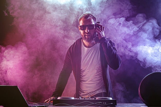 Young stylish man in glasses posing behind mixing console on colored smoke studio