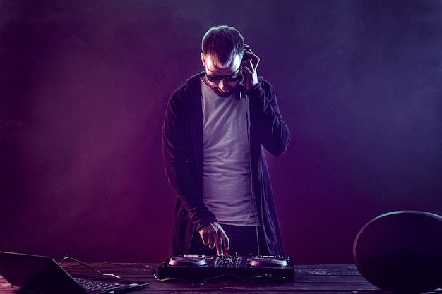 Young stylish man in glasses posing behind mixing console on colored smoke studio 