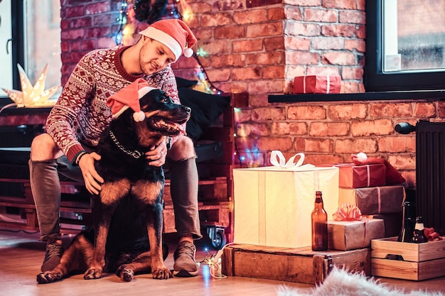 A young stylish man holding a gift box while sitting with his cute dog in a decorated living room at Christmas time.