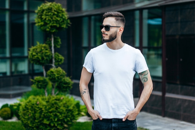 Un giovane uomo alla moda con la barba in una maglietta bianca e occhiali da sole