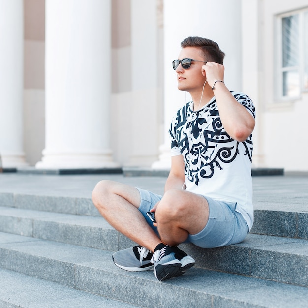 Young stylish man with sunglasses sitting on the stairs and listening to music.