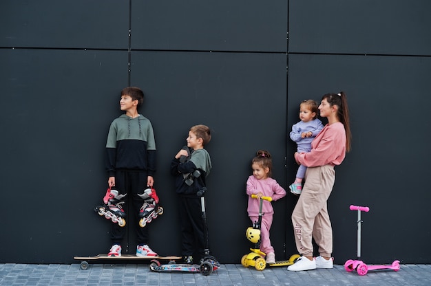 Giovane madre alla moda con quattro bambini all'aperto contro il muro moderno nero. la famiglia sportiva trascorre il tempo libero all'aperto con scooter e pattini.