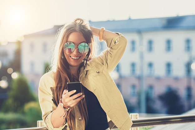 Young stylish pretty woman hands holding a phone sunset background sunny daygood weather sunglasses cool accessories