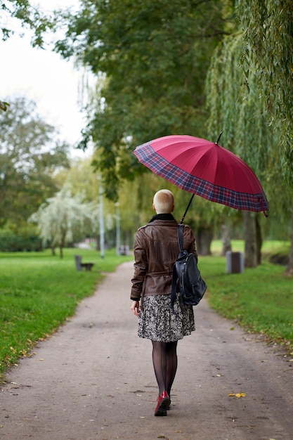 사진 젊고 세련된 짧은 머리 금발 여성이 뒤에서 빨간 우산을 들고 가을 공원을 걷는다