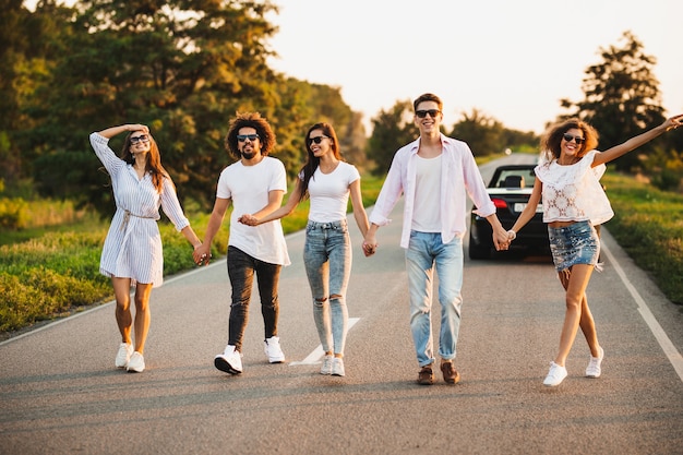 Young stylish two guys and three girls are holding hands and walking on the road on a sunny day .