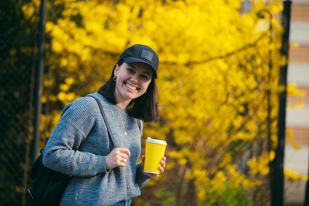 黄色い紙コップから屋外でコーヒーを飲む帽子の若いスタイリッシュな女性