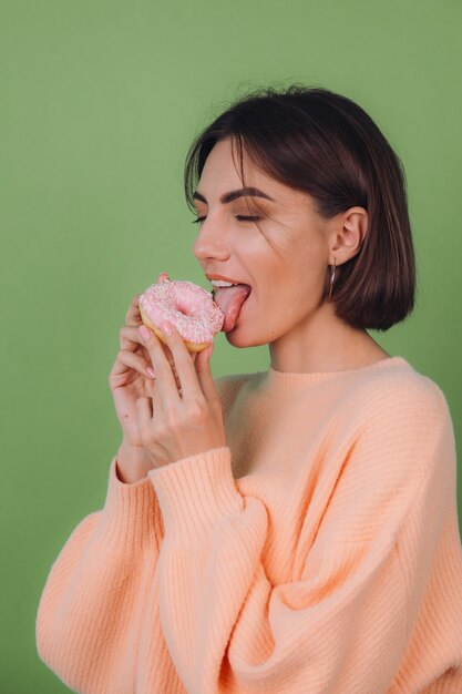 Young stylish woman in casual peach sweater  isolated on green olive wall licking pink donut copy space