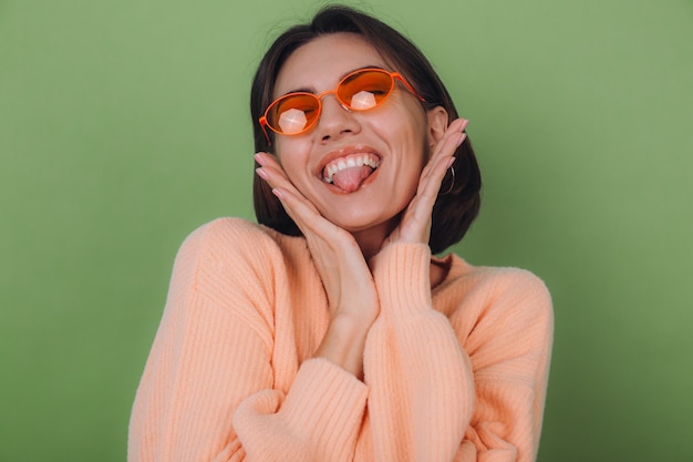 Young stylish woman in casual peach sweater and orange glasses isolated on green olive wall happy positive smiling laugh around copy space