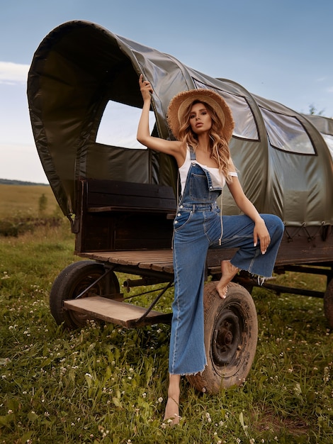 Молодая стильная женщина в джинсовом комбинезоне позирует возле старой деревянной кареты в поле