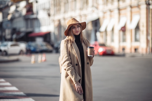 コートとフェルトの帽子をかぶったスタイリッシュな若い女性が街を歩き回り、外出先でコーヒーを飲みます。秋の時間