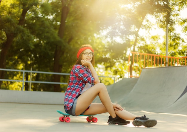 Young stylish woman dressed in youth clothes sits on a skateboard at a skatepark. Summertime, sunny bright day