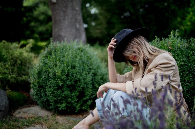 庭のラベンダーの花の近くに座っている帽子をかぶった若いスタイリッシュな女性