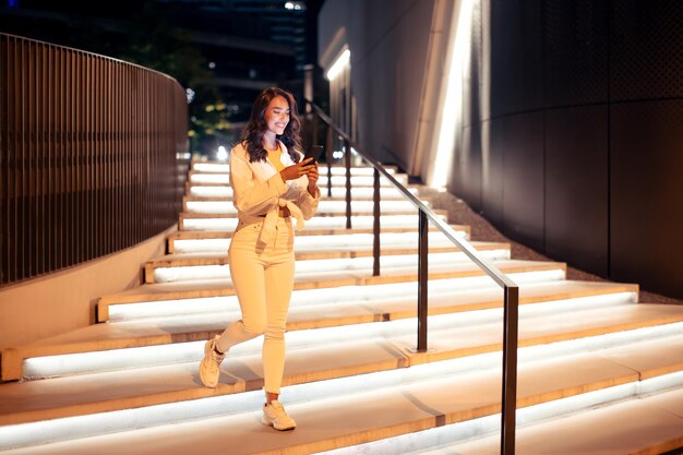 Young stylish woman holding cellphone while going down the illuminated stairs walking in the city at