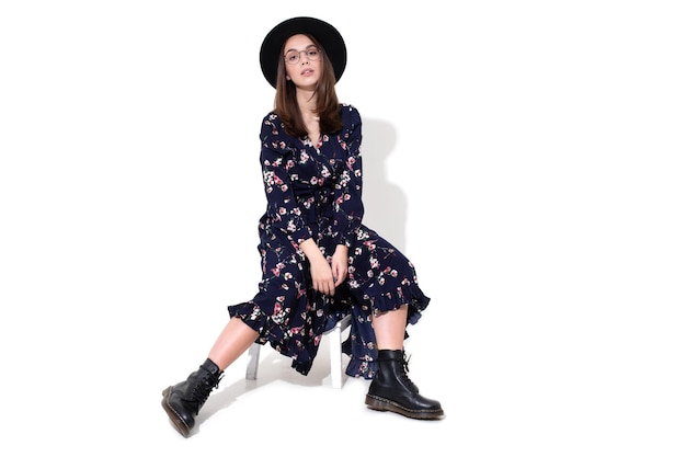 A young stylish woman is sitting on a chair in a hat and a dark dress Fashion industry photos on a white background