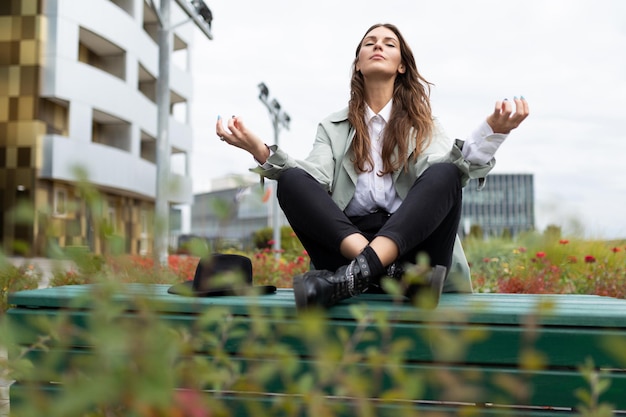 Young stylish woman meditates during a lunch break in the park against the backdrop of an office