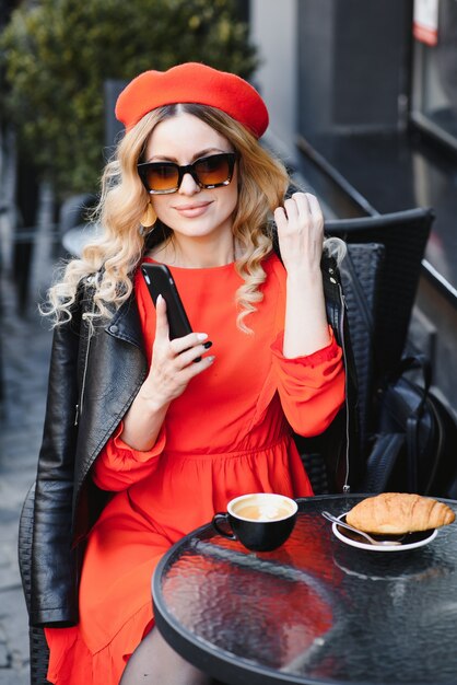 Young stylish woman in red beret having a french breakfast with coffee and croissant sitting oudoors at the cafe terrace